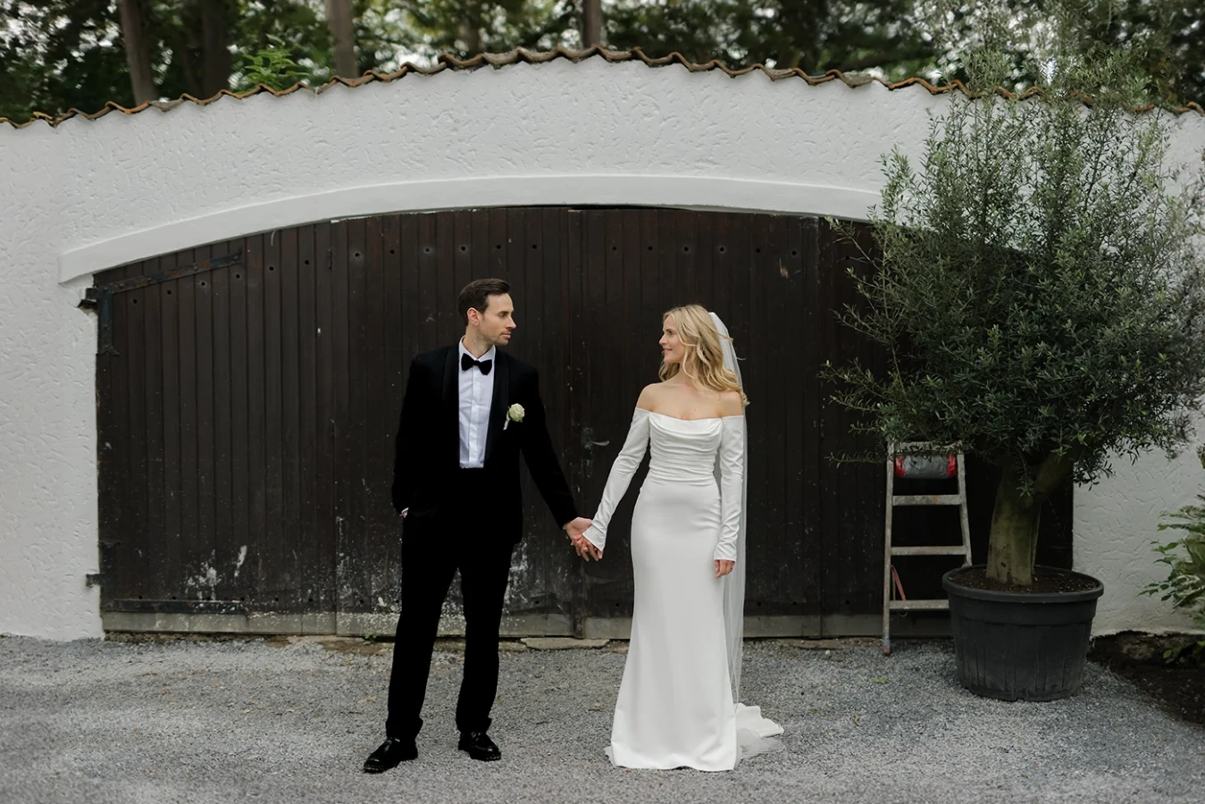 Hochzeitsfotograf Herford: Elegantes Brautpaar Hand in Hand vor rustikalem Scheunentor mit Olivenbaum, natürliche Hochzeitsfotografie